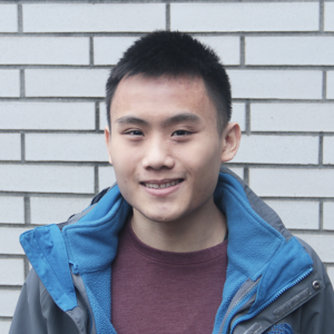 A picture of Aidan Chua smiling at the camera wearing a blue jacket and red shirt with a white brick wall.